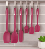 Set of pink kitchen utensils hanging on a rack with a plant in the corner.